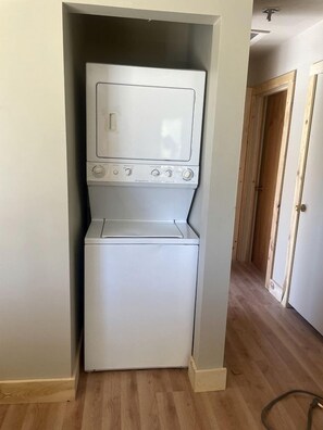 A compact laundry room with a washer and dryer, wooden flooring, and a hallway leading to a door. - Spacious Cabin with Fire Pit, Perfect for a Family Getaway to Custer State Park (Sturgis)