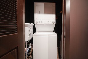 A laundry room with a stacked washer and dryer, a metal rack with clothes, and a wooden door. - The Shumaker | Unit 109 | Stylish Lancaster Apt (Lancaster)