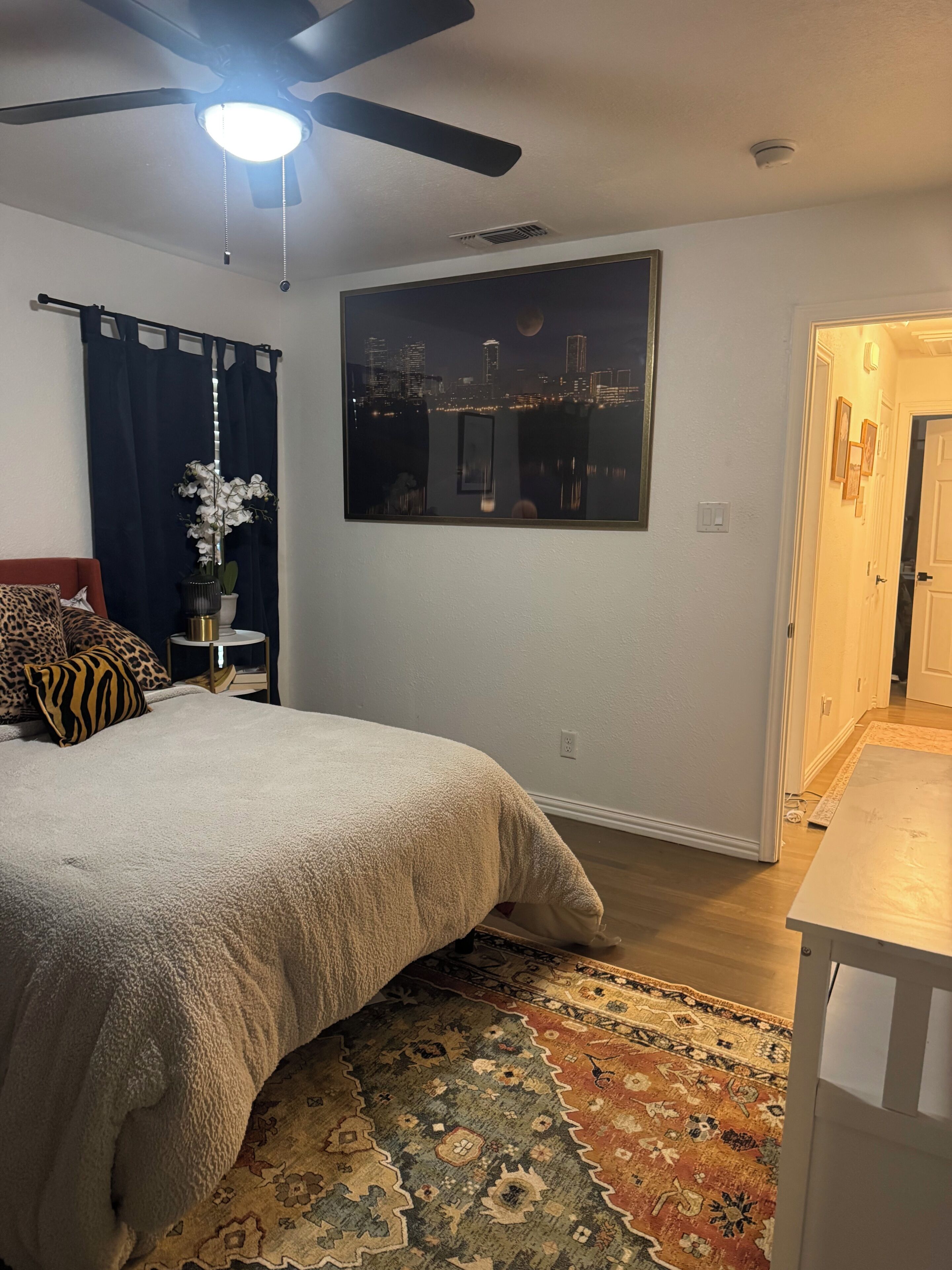 A bedroom with a bed, a ceiling fan, a nightlight, a large window with a cityscape view, a door, and a rug.