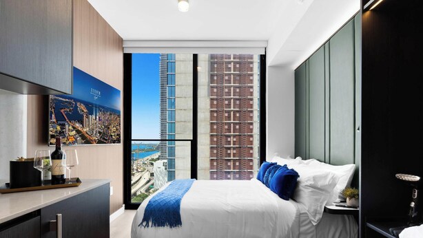 Une chambre d’hôtel moderne avec un grand lit, un bureau agrémenté d’une bouteille de vin et de verres, une vue sur le paysage urbain et un balcon donnant sur un immeuble de grande hauteur.