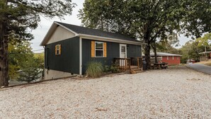 A single-story house with a dark exterior, a front porch, and a gravel driveway. - The Hideaway on Table Rock Lake (Reeds Spring)