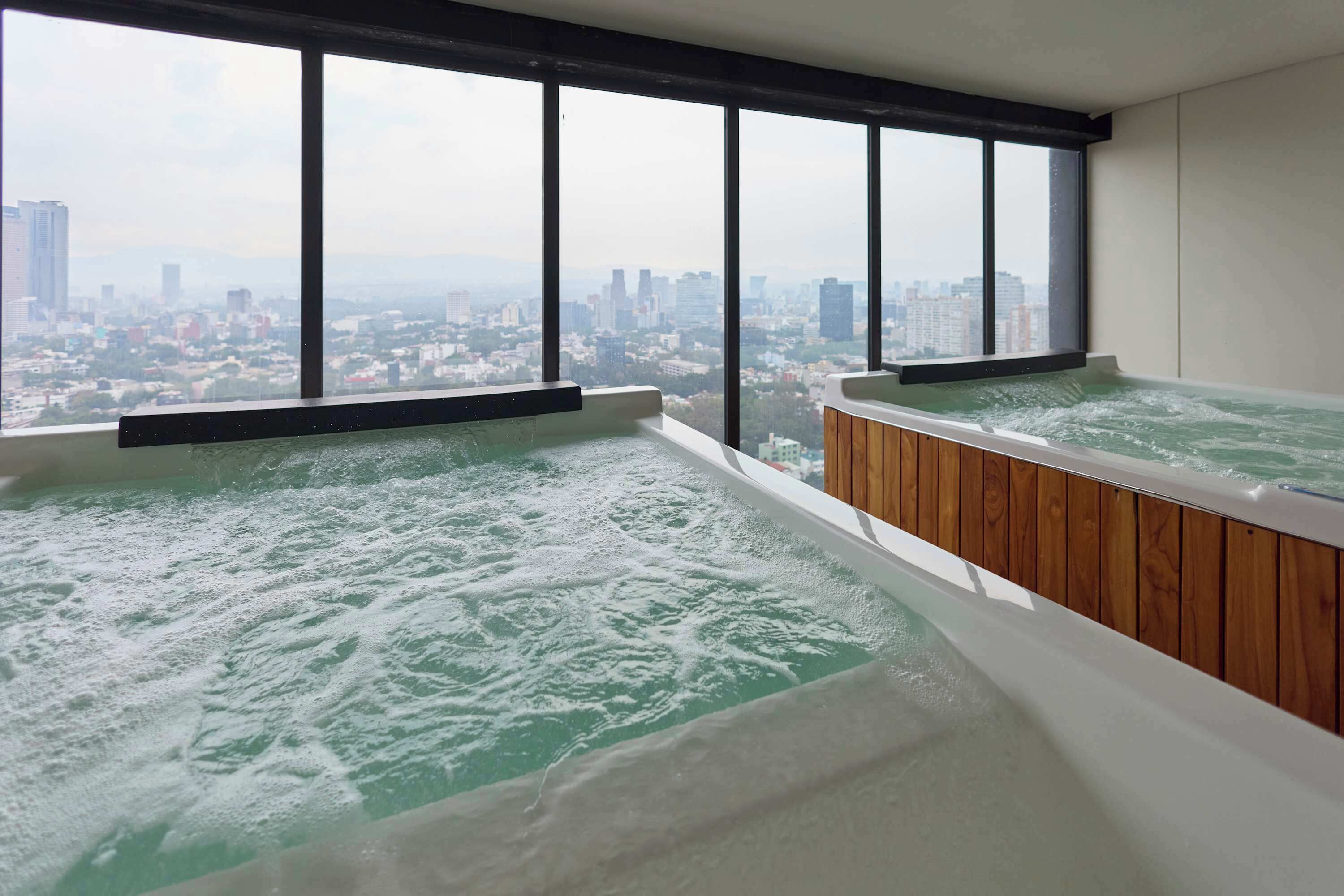 A high-rise hotel room with two hot tubs, large windows, and a cityscape view.