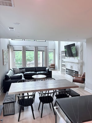 A modern living room with a sectional sofa, a dining table with chairs, a TV mounted on a marble wall, and large windows with curtains. - Modern luxury in Historic Downtown McKinney (McKinney)
