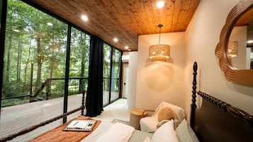 A bedroom with a wooden ceiling, a large window, a bed, a chair, a bedside table, and a mirror.