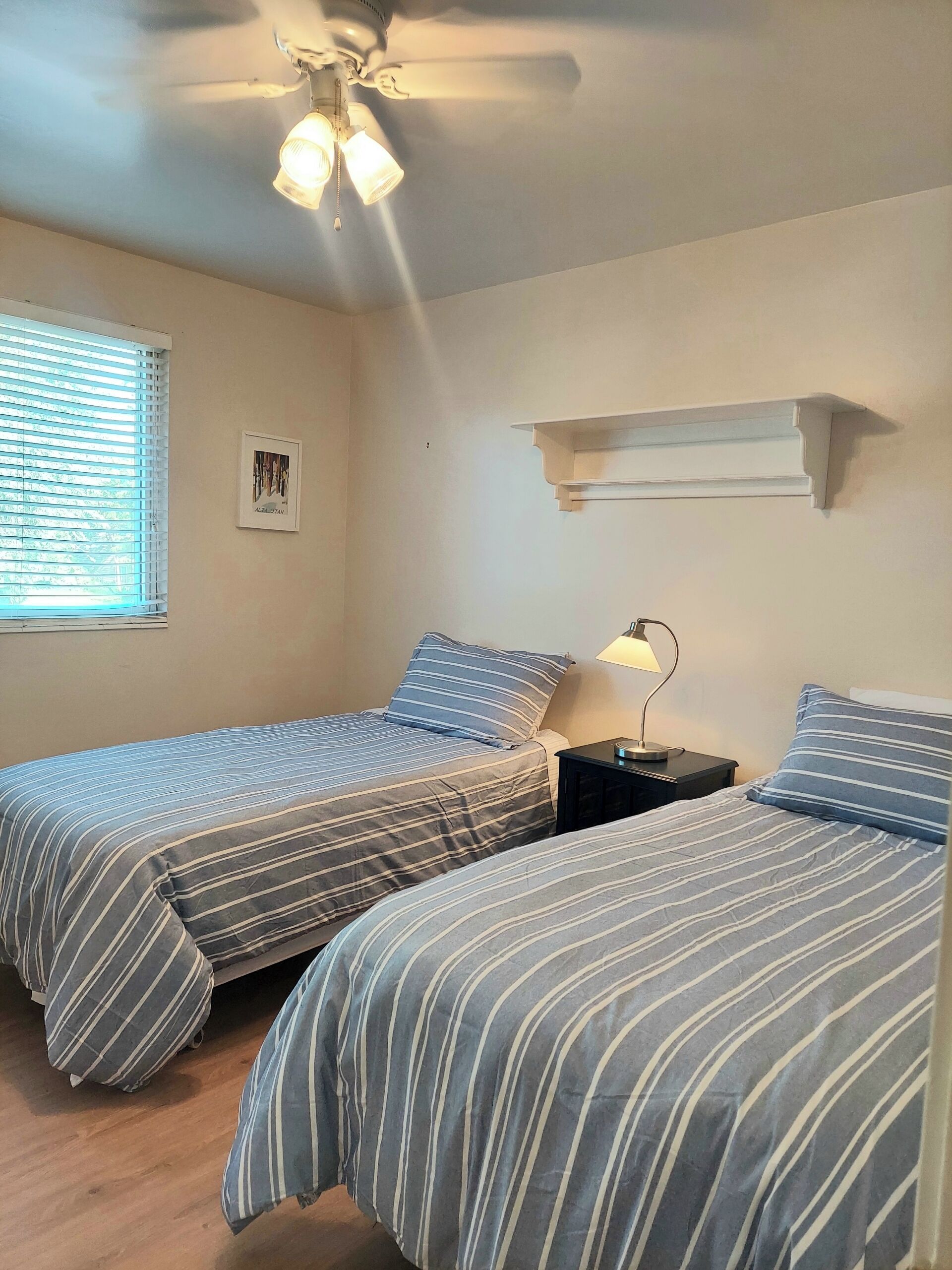 Une chambre avec deux lits, un ventilateur de plafond, une table de chevet et une fenêtre avec des stores.