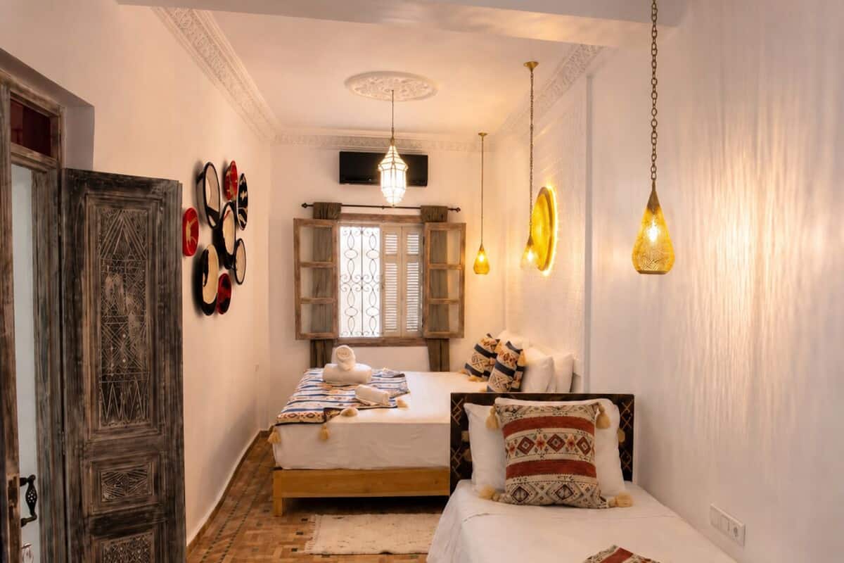 A bedroom with two beds, a wooden wardrobe, a patterned rug, and hanging lights.