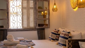 Ein Schlafzimmer mit Bett, Kissen, einem Fenster mit Rollläden, einem Spiegel und hängenden Pendelleuchten.
