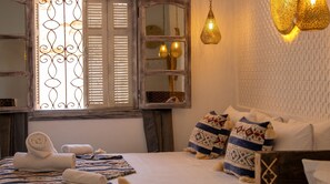 A bedroom with a bed, pillows, a window with blinds, a mirror, and hanging pendant lights. - Riad tumsilt (Marrakech)