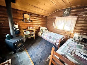 A cabin with a wood-burning stove, a bed with a quilt, a shelf with items, and a window with curtains.