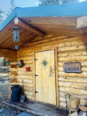 A wooden cabin with a door, a welcome sign, a bucket, and a shovel outside.