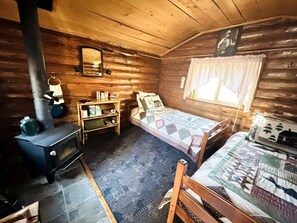 Una cabaña con estufa de leña, una cama con colcha, una repisa con objetos y una ventana con cortinas.