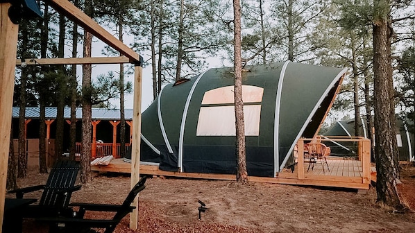 A large tent-like structure with a window, surrounded by trees, and a wooden deck with chairs.