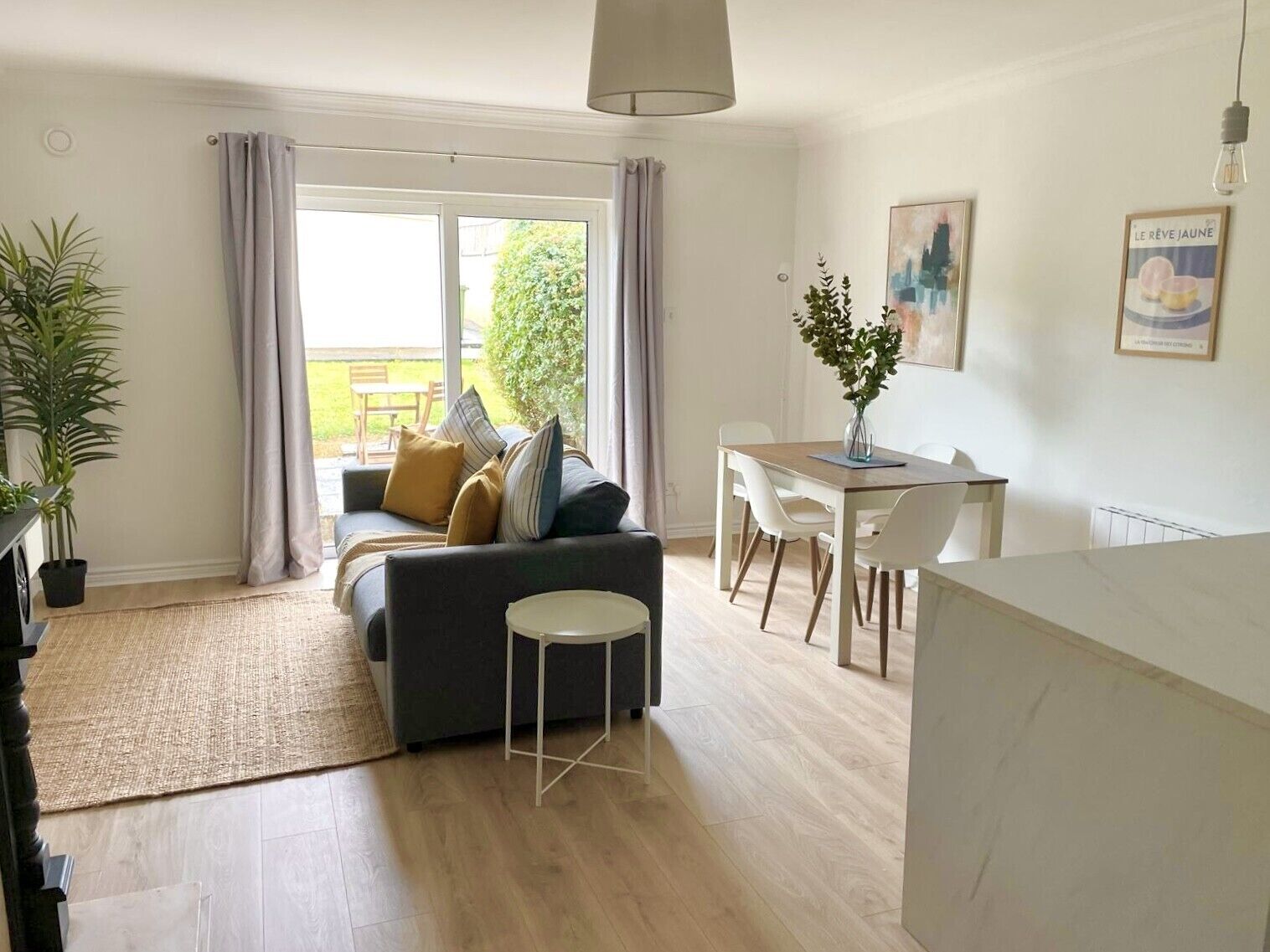 Una sala de estar moderna con sofá, mesa de comedor y sillas. Cuenta con un ventanal con cortinas, una planta en maceta y una lámpara colgante.