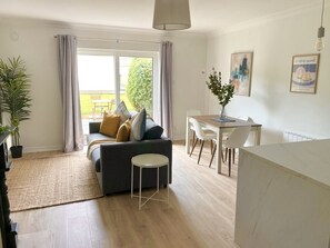 A modern living room with a sofa, dining table, and chairs. There is a large window with curtains, a potted plant, and a hanging light fixture. - Luxury By The Coast 9 (Galway)