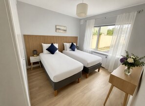 A hotel room with two beds, a bedside table with a lamp, a window with curtains, and a small table with a vase of flowers. - Luxury By The Coast 9 (Galway)