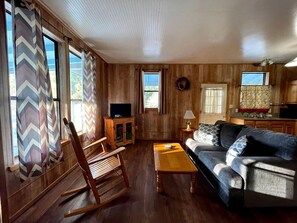 Ein Zimmer mit Holzwandverkleidung, einem Schaukelstuhl, einer Couch, einem Fernseher auf einem Schrank und einer Küchenzeile.