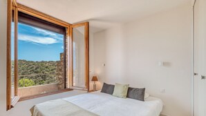 Ein Schlafzimmer mit großem Fenster, einem Bett, einer Nachttischlampe und Blick auf eine Hügellandschaft.