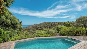 Ein rechteckiger Pool mit klarem, blauem Wasser, umgeben von einer Holzterrasse, vor einer Kulisse aus üppiger Vegetation und blauem Himmel.