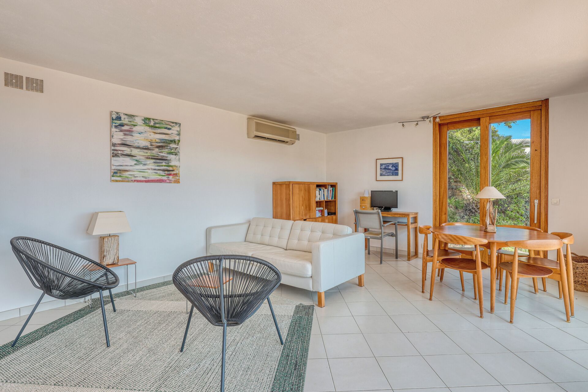 Un salon comprenant un canapé, deux fauteuils, une table à manger avec des chaises, une bibliothèque, un téléviseur et une fenêtre donnant sur un paysage verdoyant.
