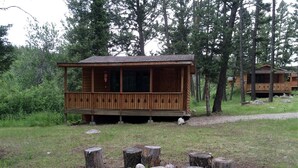 Eine Holzhütte mit Veranda, umgeben von Bäumen und einem Feuerkorb mit Holzscheiten und Steinen.