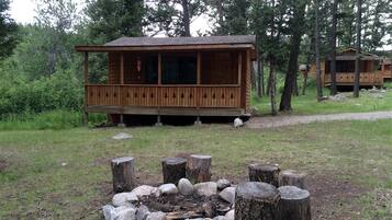 A wooden cabin with a porch, surrounded by trees and a fire pit with logs and rocks.