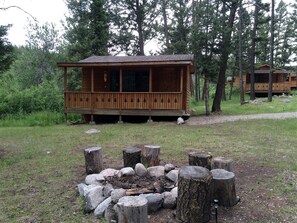 A wooden cabin with a porch, surrounded by trees and a fire pit with logs and rocks.