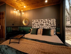 A bedroom with a bed, a chandelier, a headboard with a floral pattern, and a view of trees through a window.