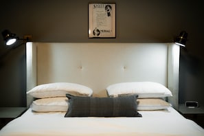 A neatly made bed with white and gray pillows, a framed safety and moral poster on the wall, and two bedside lamps. - The Shumaker | Unit 101 | Modern Lancaster Apt (Lancaster)