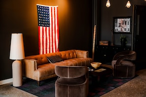 Una habitación con un sofá de cuero, dos sillones, una lámpara, una foto enmarco y una bandera estadounidense.