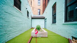 Une ruelle étroite avec un tapis d’herbe artificielle verte, un drapeau rouge et un bac à fleurs contenant des plantes.