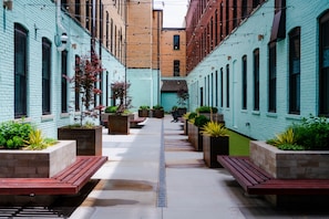 A narrow alleyway with brick buildings, benches, planters, and a central drainage grate. - The Shumaker | Unit 107 | Chic Lancaster Apt (Lancaster)