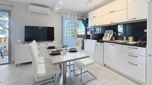 A modern kitchen with white cabinetry, a dining area with a table and chairs, and a flat-screen TV mounted on the wall. - The Oasis Family T3Clim: Terraces, Parks, and Beaches Within Walking Distance (Fréjus)