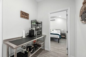 Eine kompakte Küche mit Mikrowelle, Kaffeemaschine und einer Ablage für Küchenutensilien. Ein Bett ist im angrenzenden Raum zu sehen.
