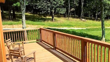 A wooden deck with a railing, chairs, and a view of a grassy area with trees.