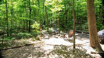 A forested campsite with several Adirondack chairs arranged around a central fire pit.