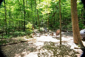 A forested campsite with several Adirondack chairs arranged around a central fire pit.