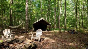 Un terrain de camping aménagé dans une forêt, avec deux fauteuils Adirondack, un hamac, un petit abri en bois et une table de pique-nique.