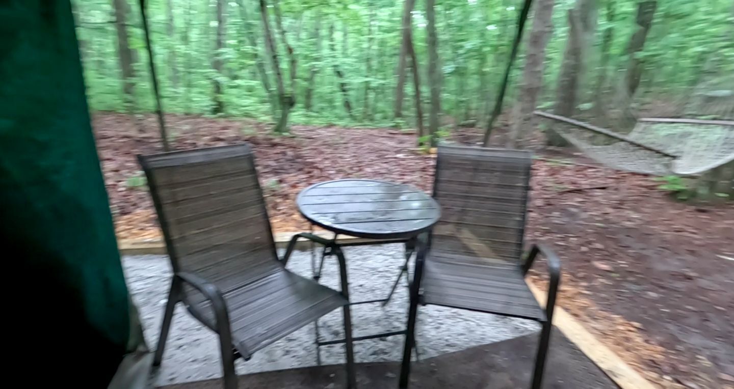 Un espace extérieur abrité, avec deux chaises et une table, entouré d’une forêt.