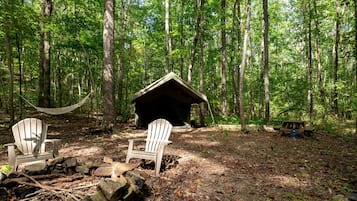 A forest campsite with two Adirondack chairs, a hammock, a small wooden shelter, and a picnic table.