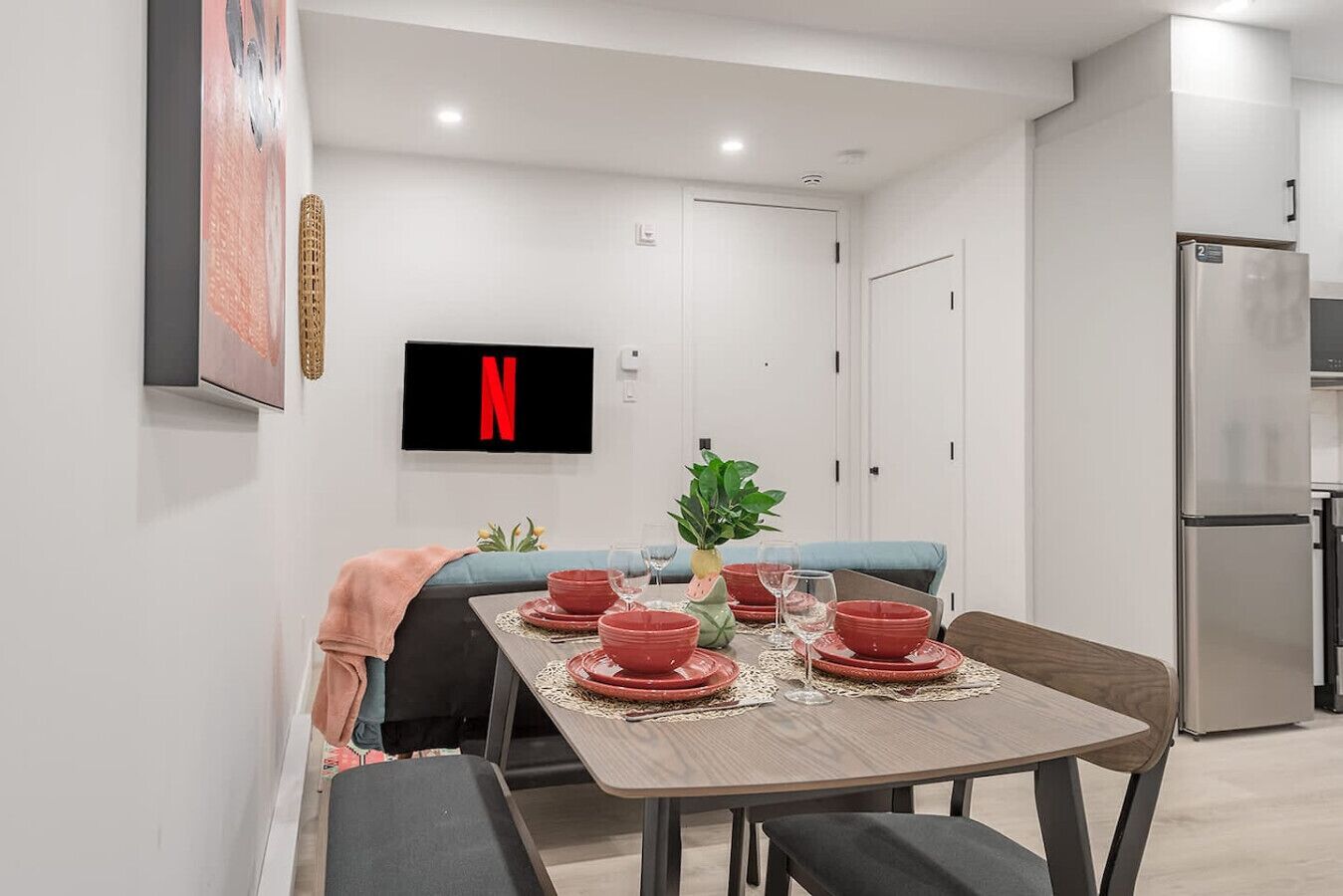Un espace repas moderne avec une table dressée pour quatre personnes, un téléviseur fixé au mur et un réfrigérateur en arrière-plan.