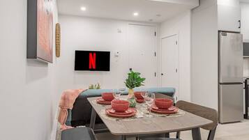 Un espace repas moderne avec une table dressée pour quatre personnes, un téléviseur fixé au mur et un réfrigérateur en arrière-plan.