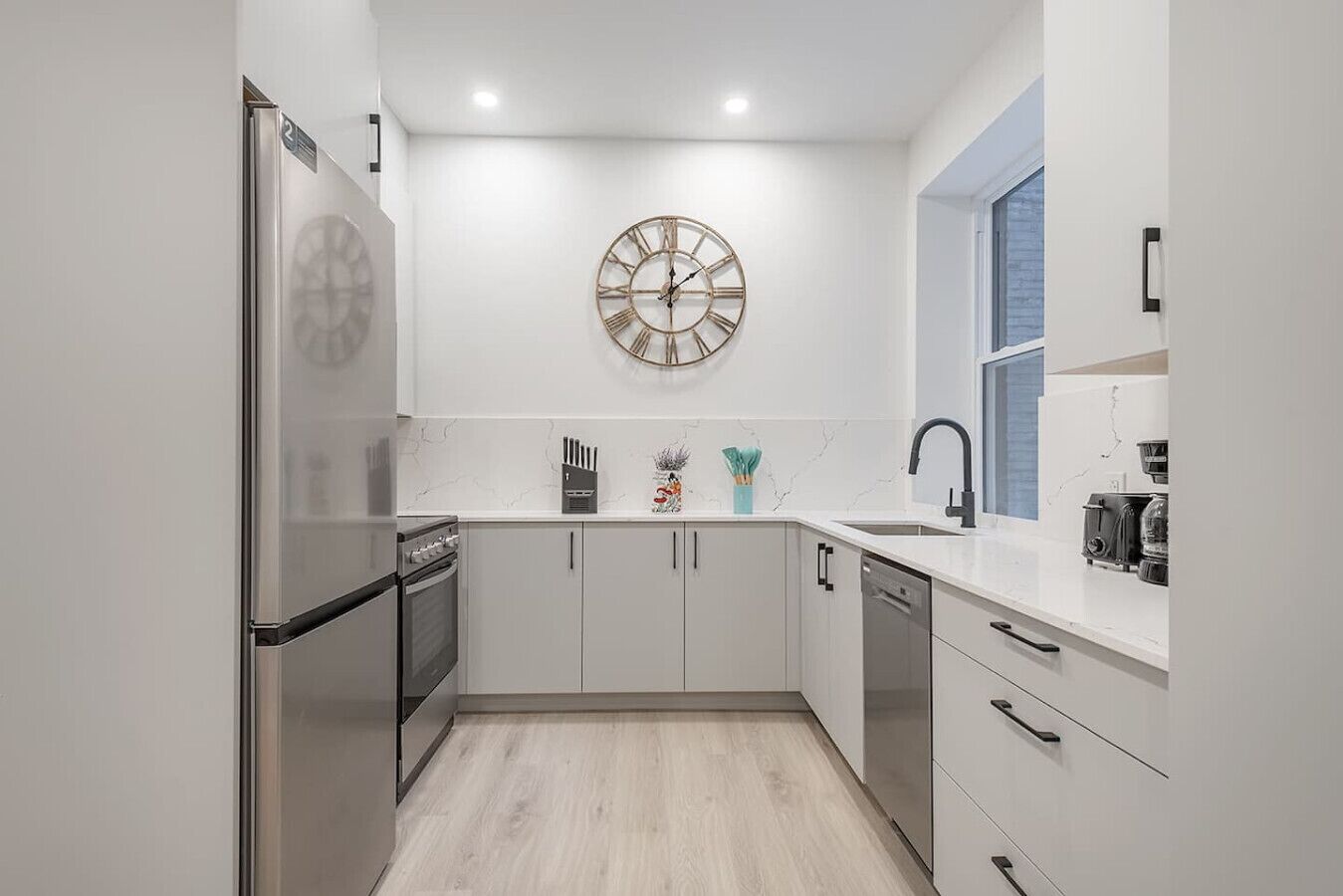 Une cuisine moderne avec des meubles blancs, un réfrigérateur en acier inoxydable, une horloge murale et une fenêtre offrant une belle vue.