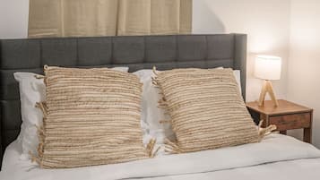 Un lit avec deux oreillers à texture, une tête de lit rembourrée de couleur grise, une table de chevet avec une lampe et une fenêtre avec des rideaux.