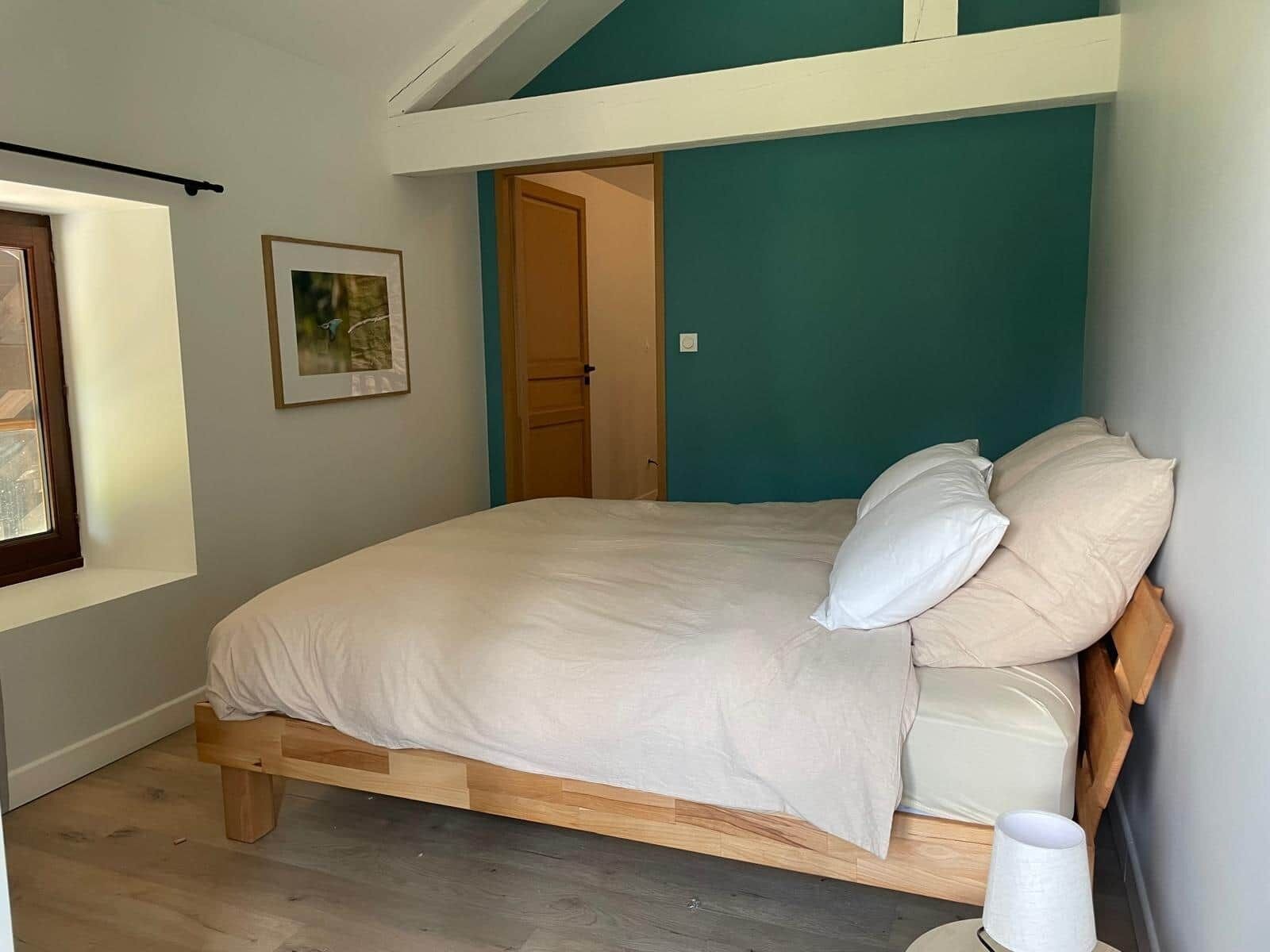 Un dormitorio con cama, ventana, puerta y un cuadro en la pared.