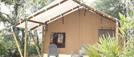 Una carpa pequeña de un piso con estructura de madera y techo de lona, dos sillas afuera y una ventana.