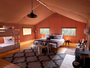 A tent-style room with bunk beds, a central bed, a desk with a chair, and a window with a view of greenery.