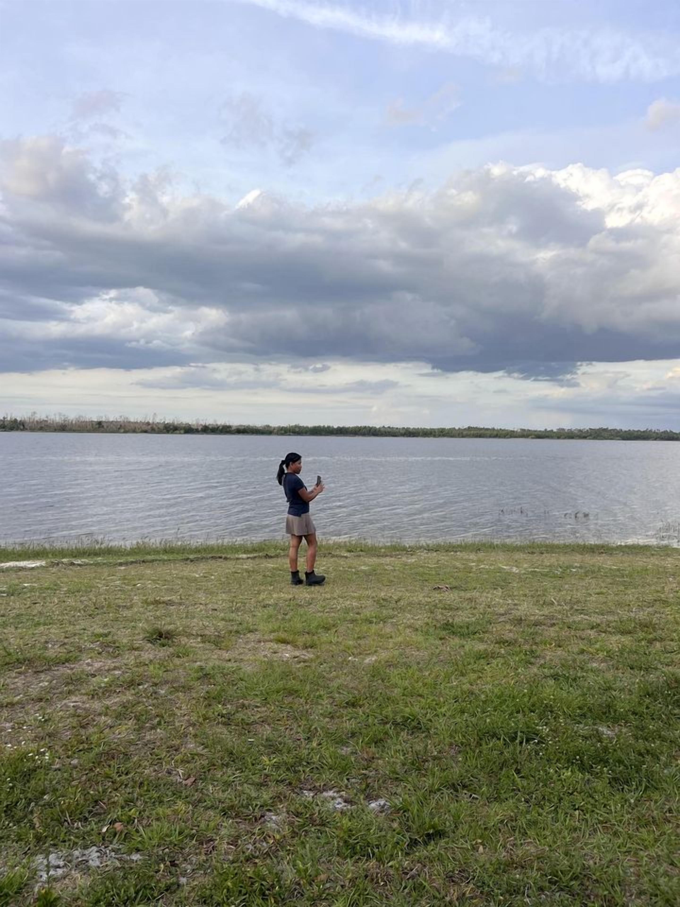 A person standing by a lake with a grassy foreground and a cloudy sky.