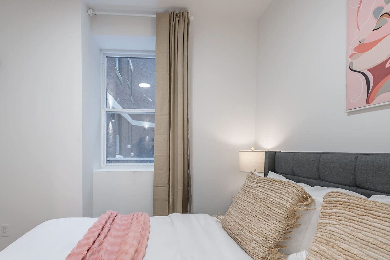 Ein Hotelzimmer mit Bett, Fenster, Lampe und einem Gemälde an der Wand.