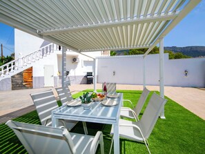 Een overdekt terras buiten met een tafel, stoelen, een barbecue en een trap naar een hoger niveau.