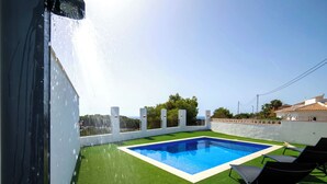 Piscina no terraço com grama sintética, uma parede branca e céu limpo.